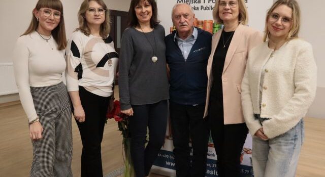 Na zdjęciu znajduję się Krzysztof Wielicki oraz pięć bibliotekarek, trzy stoją po lewej stronie, a dwie po prawej. Za nim znajduję się baner z logiem biblioteki.