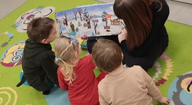 Na zdjęciu znajduję się kobieta, która trzyma otwartą książkę przedstawiającą zimowy krajobraz, obok siedzi trójka dzieci i przygląda się ilustracji. Zdjęcie zostało wykonane od góry.