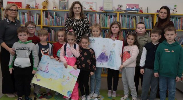Na zdjęciu znajdują się trzy Panie, wraz z dwunastoma przedszkolakami, część z nich trzyma w ręce plakaty z Krain Lodu, które same pokolorowały. W tle znajdują się półki z książkami.