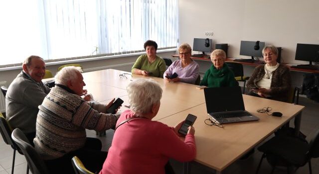 9 seniorek i seniorów siedzi przy połączonych stołach, w rękach trzymają telefony komórkowe, w tle biurka z komputerami