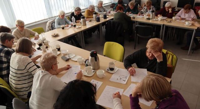 około 20 seniorek siedzi przy stołach ustawionych w literę u i pisze na kartkach, w tle biurka z komputerami