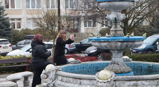 Na zdjęciu znajdują się dwie nastolatki przy fontannie, jedna robi zdjęcie. W tle znajdują się drzewa, stojące auta i budynek.