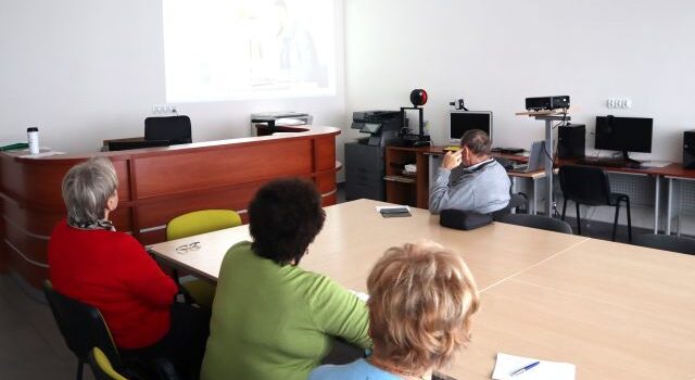 4 seniorów siedzi przy stołach i ogląda prezentację wyświetlaną na ścianie, w tle biurka z komputerami