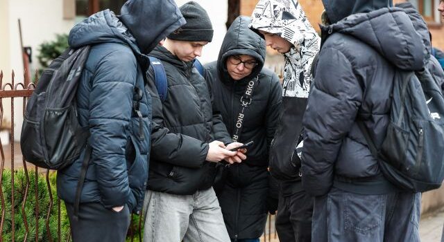 Na zdjęciu znajduje się piątka chłopaków nachylona nad telefon trzymanym przez jednego z nich. Chłopacy znajdują się na dworze.