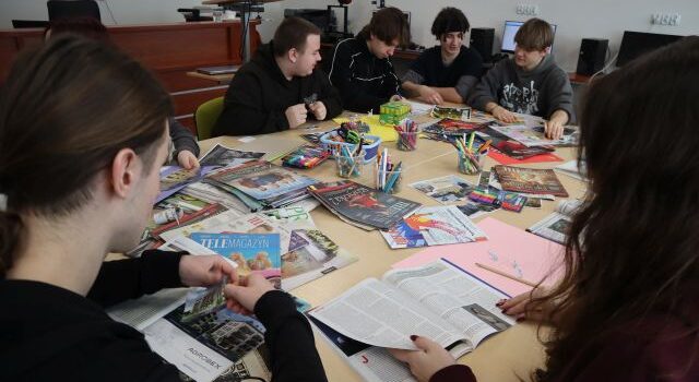 Na zdjęciu znajduja się uczniowie przy złączonych stołach, na których znajdują sie kartki i gazety, z których wykonują kolaże. Warsztaty odbywają się w sali komputerowej.
