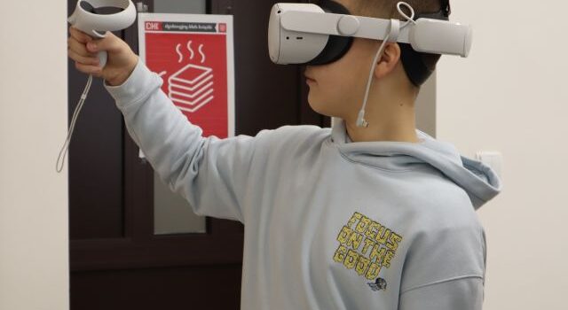 Na zdjęciu znajduje się chłopiec bokiem w okularach VR, który w uniesionej ręce na poziomie twarzy trzyma kontroler. W tle znajdują się drzwi z plakatem.