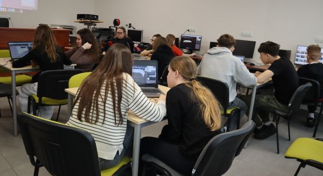Na zdjęciu znajduje się młodzież przy stolikach z laptopami oraz przy komputerach i tworzą swoją wirtualną galerię. Warsztaty odbywają się w sali komputerowej.