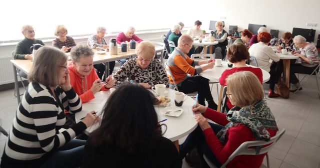 Na zdjęciu znajdują się kobiety przy stolikach, kobiety z tyłu siedzą przy złączonych kwadratowych stołach, a kobiety na przodzie siedzą przy trzech okrągłych stołach, na których znajduje się kawa, herbata i ciasteczka. Zajęcia odbywają się w sali komputerowej.