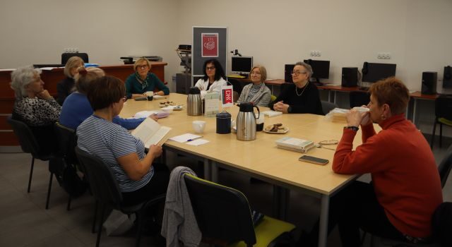 Na zdjęciu przy złączonych stolikach siedzi dziewięć kobiet, jedna z nich ma w ręce kartkę. Na stole znajdują się termosy, szklanki i ciasteczka. Zajęcia odbywają się w sali komputerowej.