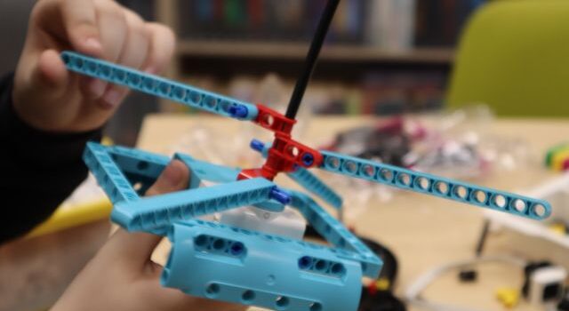 Na zdjęciu znajduje się czubek helikoptera zbudowanego z klocków LEGO, konstrukcja jezt trzymana przez uczestnika zajęć, na zdjęciu uchwycone zostały tylko ręce. W tle znajduje się stół, na którym leżą klocki, kawałek krzesła oraz półki z książkami.