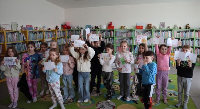 Na zdjęciu znajduje się siedamnaście przedszkolaków stojących do zdjęcia ze swoimi pracami. W tle znajdują się półki z książkami.