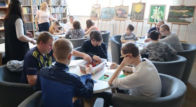 Na zdjęciu znajduje się młodzież siedząca przy okrągłych stołach, przy których układają klocki LEGO. W tle znajdują się obrazy oraz okna, a po prawej stronie półki z książkami.