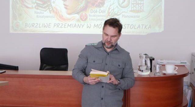 Na zdjęciu znajduje się mężczyzna z książką w ręku, który opiera się o biurko, w tle znajduje się wyświetlana na ścianie prezentacja.