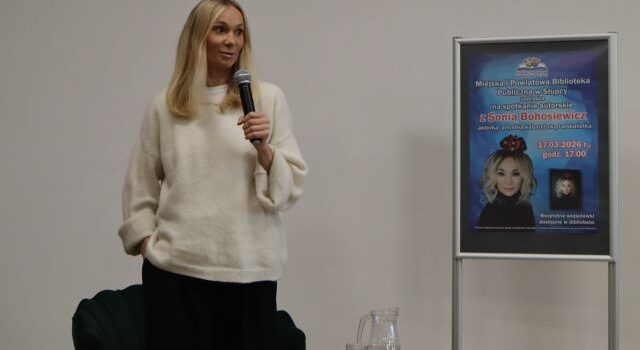 Na zdjęciu znajduje się kobieta stojąca na scenie z mikrofonem, obok niej stoi plakat promujący spotkanie.