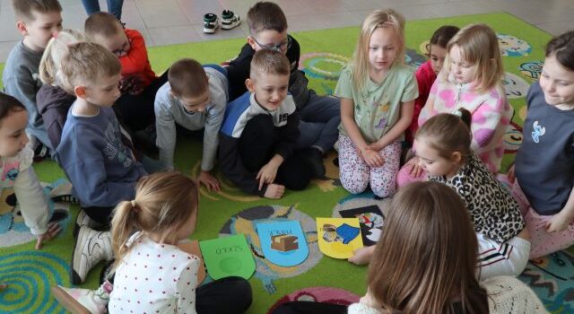 Na zdjęciu znajduje się grupa przedszkolaków na dywanie wraz z kobietą, na środku znajdują się wycięte z papieru kolorowe kosze.