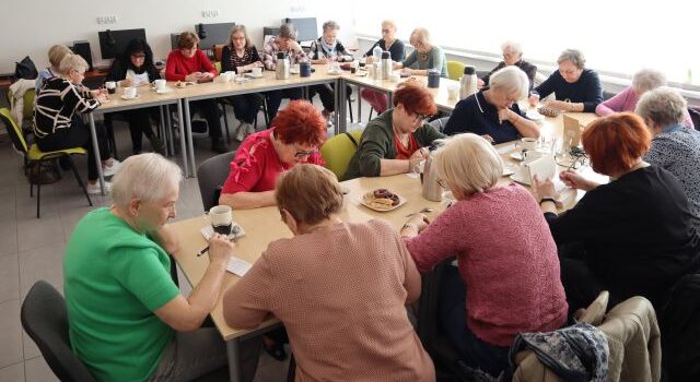 Na zdjęciu znajduje się dwadzieścia kobiet, które siedzą przy załączonych w literę C stolikach i rozwiązują zadania przy kawie. Warsztaty odbywają się w sali komputerowej.