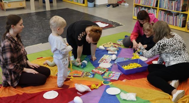 Na zdjęciu znajduje się trójka dzieci oraz cztery kobiety na kolorowej chuście, na której znajdują się książeczki, papierowe talerzyki. W tle znajdują się półki z książkami, a na podłodze leży folia.