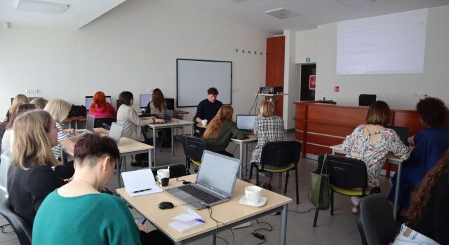 Na zdjęciu znajdują się kobiety oraz jeden mężczyzna przy stolikach z laptopami oraz przy trzech komputerach. Po prawej stronie na ścianie wyświetlana jest prezentacja o sztucznej inteligencji. Warsztaty odbywają się w sali komputerowej.