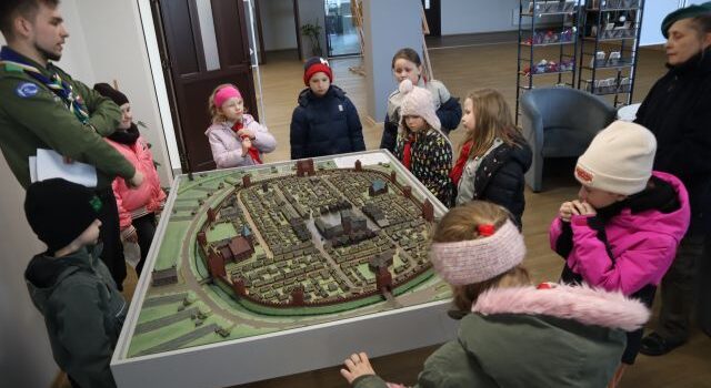 Na zdjęciu znajduje się grupka dzieci oraz dwóch harcerzy i stoją dookoła makiety miasta, która znajduje się na holu biblioteki.