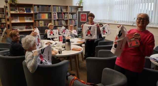 Na zdjęciu znajdują się kobiety na fotelach przy stolikach, dwie z nich stoją i wszystkie trzymają w ręce upominki w postaci białych torb z nadrukiem. W tle znajdują się półki z książkami oraz okna.