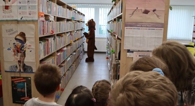 Na zdjęciu znajdują się dzieci przy półkach z książkami, pomiędzy którymi stoi człowiek w przebraniu niedźwiedzia.