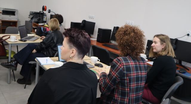Na zdjęciu znajdują się trzy kobiety przy stoliku, na którym znajduje się laptop, przed nimi pry stoliku siedzą dwie kobiety. Warsztaty odbywają się w sali komputerowej.