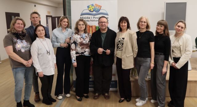 Na zdjęciu się dwóch mężczyzn oraz osiem kobiet stojących wspólnie do zdjęcia na holu biblioteki, za nimi stoi baner z logiem biblioteki.