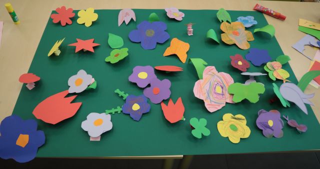 Na zdjęciu znajduje się zielony brystol z kolorowymi kwiatkami, przedstawiający łąkę. Praca znajduje się na złączonych drewnianych stolikach.