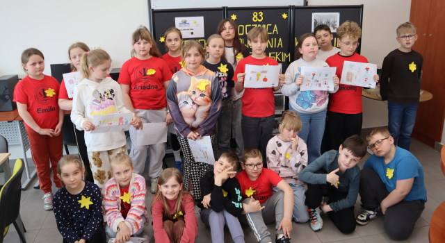 Na zdjęciu znajduje się dwudziestu jeden uczniów, część ma w ręce kartki i przyklejone na koszulce żółte żonkile. Uczniowie stoją do zdjęcia na tle czarnej tablicy upamiętniającej 83 rocznice wybuchu powstania w getcie warszawskim.